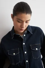 Camisa de jean de corte recto y calce holgado, con cuello clásico, cierre frontal con botones metálicos y dos bolsillos delanteros con solapa y botón.