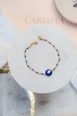 Pulsera de cadena fina dorada con pequeñas cuentas azules intercaladas y un dije central de ojo turco en color azul y blanco. Cuenta con cierre de mosquetón y cadena de extensión.