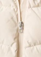 Campera acolchada de corte recto y diseño puffer, color beige, con capucha, cuello alto y cierre frontal de cremallera doble. Cuenta con mangas largas con puños elásticos y dos bolsillos laterales con cierre.
