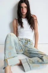 Jeans de tiro bajo confeccionados en denim, con diseño holgado y estampado integral de rosas en tonos amarillos y verdes sobre fondo celeste claro. Cuenta con cinco bolsillos, trabillas para cinto y botamangas con vuelta.