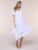 Vestido midi blanco de estilo romántico confeccionado en poliéster con elastano, con detalles de encaje floral. Cuenta con escote cruzado en V, mangas cortas acampanadas, cintura elastizada y falda evasé con terminación asimétrica en picos.