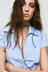 Camisa de color celeste con cuello solapa, escote en pico y manga corta. Presenta un diseño entallado con detalle de frunces verticales en la parte central y cierre frontal con botones.