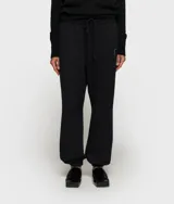 Pantalón deportivo negro de corte holgado y largo, confeccionado en algodón con elastano. Presenta cintura elástica con cordón ajustable, puños elásticos en los tobillos y un pequeño parche con un logo en forma de contorno de figura humana en la cadera izquierda.