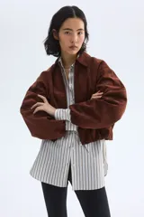 Campera corta marrón de tejido suave con terminación cepillada, cuello camisero, hombros caídos y cierre frontal. Cuenta con bolsillos laterales ribeteados y elástico revestido en puños y dobladillo.