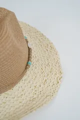 Sombrero de paja de ala ancha, con copa estilo fedora en color marrón y ala en color beige claro con hilos dorados entretejidos. Presenta una cinta decorativa con cuentas turquesas y un lazo de cordón fino atado alrededor de la base de la copa.