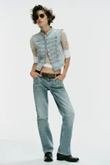Chaleco de denim color celeste con cuello subido, sin mangas, con detalle de aplicaciones horizontales y botones metálicos en el frente. Cuenta con cierre frontal con ganchos metálicos ocultos.