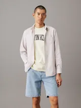 Camisa de manga larga Calvin Klein confeccionada en popelina de algodón elástico con diseño de rayas verticales. Presenta cuello en punta, cierre frontal de botones, bolsillo de parche en el pecho y bajo redondeado.