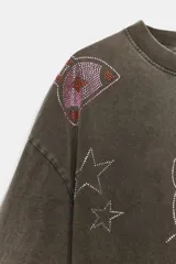 Remera oversize de color marrón grisáceo con efecto lavado, cuello redondo y manga corta. Cuenta con múltiples aplicaciones de brillos con motivos de Hello Kitty, estrellas, cerezas y moños.