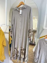 Vestido camisero largo de lino blanco con botones, cuello clásico y mangas largas con puños estampados con animal print. Tiene un corte irregular con el frente más corto que la parte trasera y estampado animal print en el ruedo.