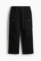 Pantalón cargo negro de corte holgado, confeccionado en tejido de algodón. Presenta cintura con elástico, bolsillos laterales al bies, bolsillos de fuelle con botones a presión en las piernas y pliegues en las rodillas.