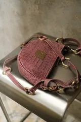 Cartera de mano con diseño semicircular, confeccionada en cuero con textura tipo crocco color negro. Presenta una placa metálica dorada con logo en el frente y un sistema de correa ajustable con eslabones y ganchos metálicos dorados que permiten variar su longitud.
