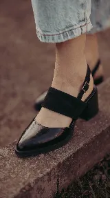 Zapato de taco ancho y medio, color negro, confeccionado en cuero. Presenta diseño destalonado con tira ajustable en el talón, faja ancha de gamuza sobre el empeine y puntera fina de charol.