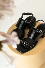 Sandalias de cuero charol negro con taco cuadrado bajo, diseño de dos tiras anchas decoradas con ojales metálicos y tira trasera ajustable con hebilla.