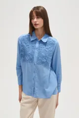Camisa de manga larga color celeste, con cuello camisero y cierre frontal de botones. Presenta un diseño distintivo con paneles de encaje tipo crochet en la parte superior delantera.