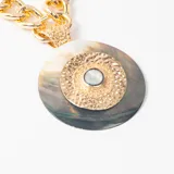 Collar de cadena gruesa de eslabones dorados con un dije circular grande que combina nácar oscuro y una pieza central texturizada en metal dorado con una piedra central.