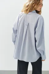 Camisa oversize de manga larga con cuello solapa, cierre frontal de botones y bolsillo de parche en el pecho, con diseño de rayas verticales celestes y blancas.
