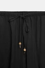 Pantalón fluido de tiro alto color verde militar, con cintura elástica ajustable con cordones y detalles de cuentas de madera. Tiene bolsillos delanteros y pierna recta.