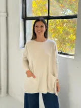 Sweater de punto color beige, de corte holgado y diseño oversize. Presenta cuello redondo, mangas largas con puños ajustados y dos bolsillos frontales tipo parche. El diseño incluye una costura vertical central en el frente y terminaciones acanaladas en el ruedo.