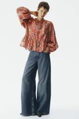 Blusa de viscosa con estampado paisley en tonos rojizos, cuello redondo con escote en V y tiras finas con cuentas decorativas. Cuenta con mangas globo largas terminadas en elástico fino y detalle de peplum acampanado en el ruedo.