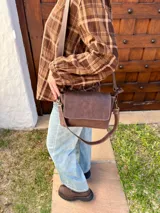 Cartera bandolera de cuero color chocolate con solapa frontal, costuras a la vista y herrajes metálicos. Incluye dos correas desmontables, una corta y una larga, y cuenta con bolsillo interior.