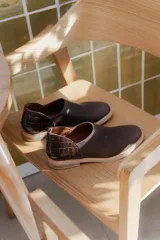 Zapatos tipo mocasín de cuero color marrón oscuro, con diseño de textura lisa en la parte delantera y textura de reptil en el talón. Cuentan con cierre trasero y suela plana de color claro.