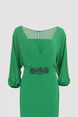 Vestido largo color verde esmeralda, con escote cuadrado y mangas 3/4 abullonadas. Presenta un cinturón con apliques de flores y una falda con paneles de gasa y volados asimétricos.