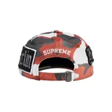 Gorra de béisbol con diseño de camuflaje en tonos rojos, blancos y negros, decorada con múltiples parches bordados que incluyen nombres de ciudades como Manhattan, London, Shibuya y Tokyo, además de la marca Supreme.