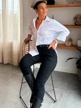 Camisa blanca de corte clásico y diseño holgado, con cuello camisero que presenta un detalle de pedrería o brillo sutil en los bordes. Posee mangas largas con puños rectos y un largo asimétrico, siendo más corta en la parte delantera y más larga en la parte trasera.