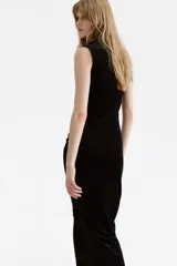 Vestido largo sin mangas confeccionado en velour suave. Presenta un diseño entallado con cuello alto y un efecto drapeado creado mediante pliegues estratégicos en el hombro y en el lateral de la cintura.