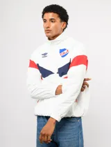 Buzo deportivo de nylon con medio cierre frontal y cuello alto. Presenta un diseño de bloques de color en azul, blanco y bordó, con el escudo del Club Nacional de Football y el logo de Umbro en el pecho.