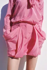 Blusa fluida de color rosado con cuello redondo ajustable mediante lazadas del mismo tejido, manga larga con puño y bajo asimétrico con aberturas laterales.