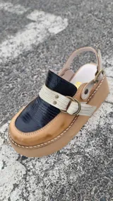 Zueco de cuero color marrón claro, con capellada en cuero negro con textura croco, tiras cruzadas frontales en cuero texturizado claro con hebilla metálica circular y tira ajustable en el talón. Cuenta con suela de plataforma corrida.
