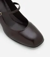 Zapato estilo Mary Jane de poliuretano color marrón oscuro, con punta redondeada, tira con hebilla ajustable sobre el empeine y taco bloque de altura media con acabado símil madera.