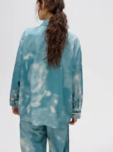 Camisa oversize de viscosa con estampado tipo tie-dye en tonos azulados. Presenta cuello clásico, cierre frontal con botones, bolsillo frontal con bordado y detalles de vivos en contraste en la tapeta y puños.