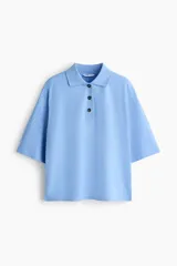 Remera tipo polo de algodón en color celeste, con diseño holgado, hombros caídos y mangas cortas que llegan hasta el codo. Cuenta con cuello acanalado y cierre frontal parcial con tres botones oscuros.