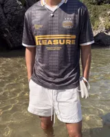 Camiseta de fútbol estilo retro de manga corta, color negro con diseño texturizado a rayas verticales, cuello en V blanco con detalle en mostaza y bordes de mangas a juego. Presenta estampados gráficos en el frente con la inscripción "PLEASURE" y emblemas deportivos.