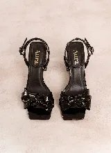 Sandalia de tacón alto y ancho, color negro charol, con punta cuadrada, tira ajustable al tobillo y detalle de moño trenzado en la parte delantera.