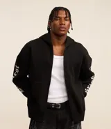 Saco tipo hoodie con cierre frontal de cremallera, color negro, corte boxy y capucha. Presenta estampa de letras en blanco en ambas mangas y un colgante de cruz metálico en el tirador de la cremallera principal.