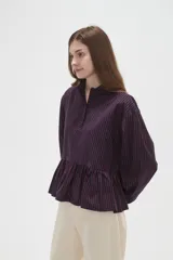 Camisa de manga larga con estampado de rayas verticales en azul marino y rojo. Presenta cuello camisero, cierre frontal con botones y un diseño con volado en la cintura que aporta volumen.