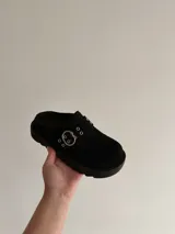 Sueco de plataforma color negro, confeccionado en material sintético tipo gamuza. Presenta una tira frontal con hebilla metálica y ojales decorativos, con suela dentada de goma.