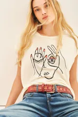 Remera de viscosa color crema, de calce suelto, escote redondo y manga corta. Presenta una estampa frontal centrada con el dibujo de un rostro y manos, y un bordado de un ojo con pedrería.