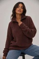 Sweater de punto grueso color marrón, con diseño de trenzas y textura, cuello alto con cierre metálico frontal y calce holgado.