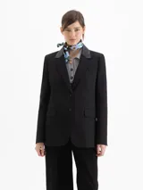 Blazer negro de corte sastrero con solapas en cuero vacuno. Presenta cierre frontal con broches forrados, bolsillos laterales con tapa y bolsillo relojero. Cuenta con tajos en la espalda y un largo que sobrepasa la cadera.