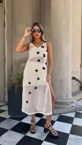 Vestido largo de tejido de red blanco, con apliques de flores negras en relieve. Lleva tirantes finos y tiene un corte recto con abertura lateral. Se usa sobre una prenda interior blanca.