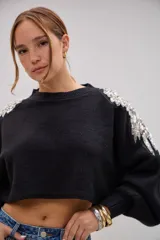 Sweater negro de corte cropped con detalle de apliques bordados en pedrería y lentejuelas plateadas sobre el hombro. Presenta mangas largas con puños ajustados y cuello redondo.