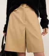 Bermuda color beige de corte recto confeccionada en tejido liso. Lleva una cinturilla transtemporal con dos botones y solapas plisadas justo debajo de la cintura.