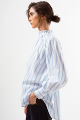 Blusa de algodón blanco con diseño de rayas verticales y textura de pequeños puntos en relieve. Presenta cuello alto con volados, cierre frontal con botones forrados y mangas largas abullonadas con puños ajustados.