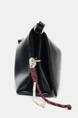 Bolso de hombro tipo baguette en color negro, con acabado brillante y estructura rectangular. Cuenta con una correa combinada de cadena metálica plateada y cuero sintético, y un pequeño monedero extraíble unido a un lateral. Presenta detalles de cordones decorativos en el otro lateral y cierre mediante solapa con imán.