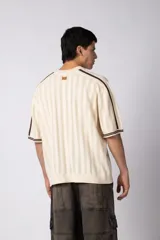 Remera de tejido de punto con textura calada, color crema con rayas verticales sutiles. Presenta cuello en V, mangas cortas con franjas laterales en color marrón oscuro y logo bordado en el centro del pecho.