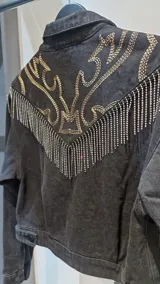 Campera de jean negra, corte a la cintura, con bordados decorativos, tachas plateadas y flecos de cadenas en la parte frontal y en las mangas.